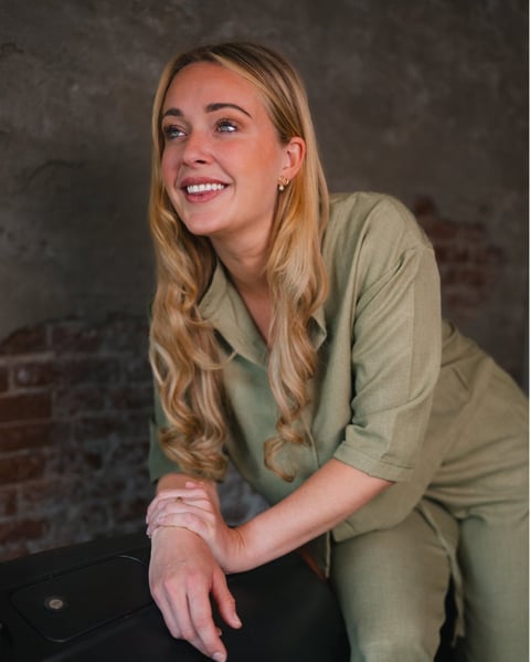 Woman with blonde wavy hair smiling while wearing a sage green button-up shirt, posed against a textured dark background