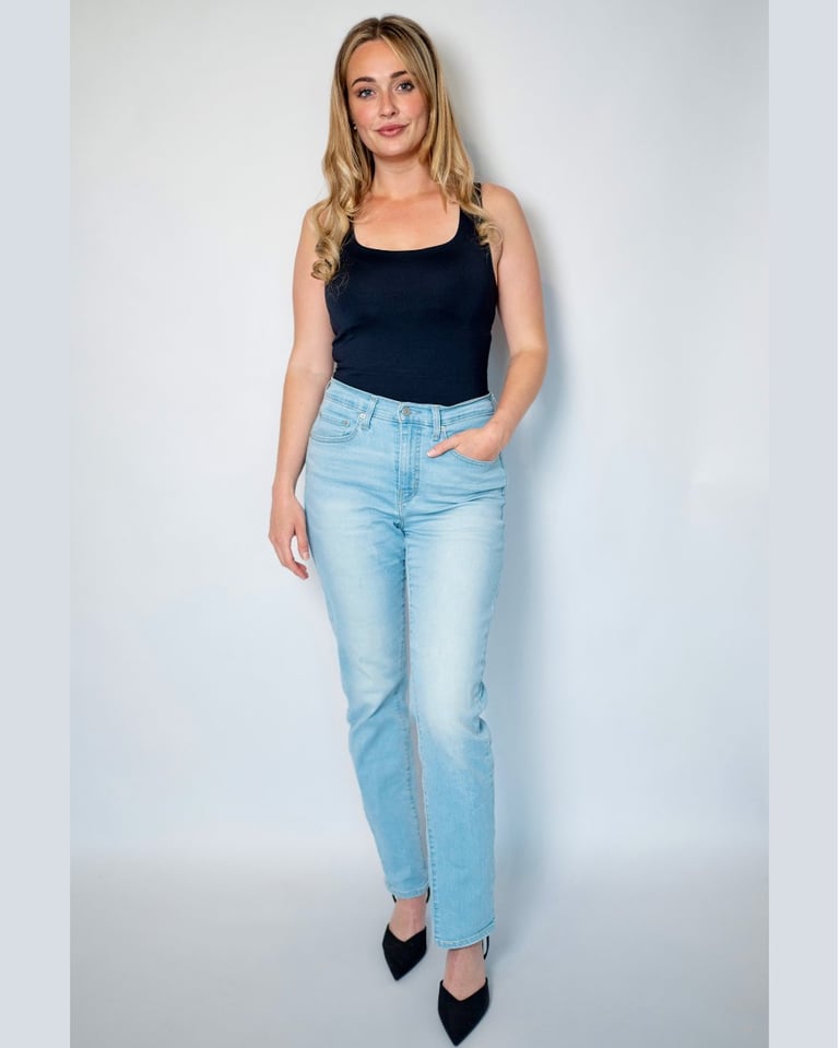 Woman wearing a black sleeveless tank top, light blue jeans, and black pointed heels against a white background