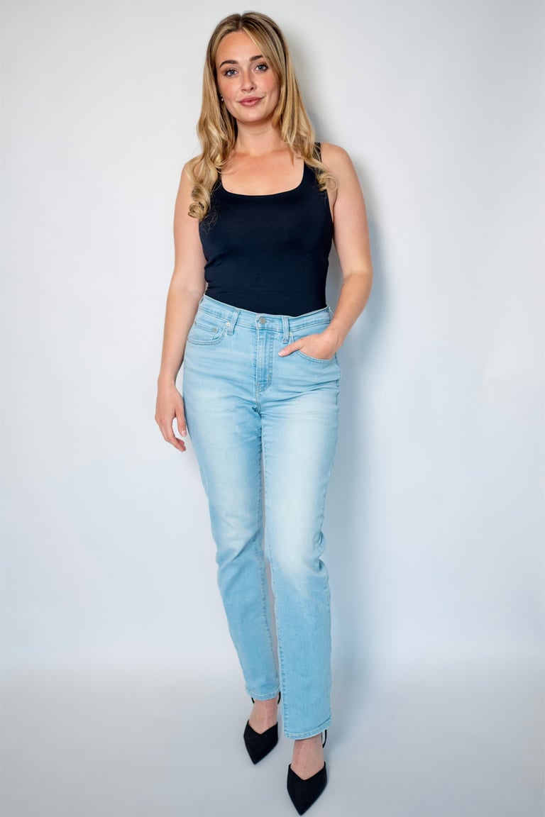 Woman wearing a black sleeveless top and light blue jeans with black pointed-toe heels, standing against a white background